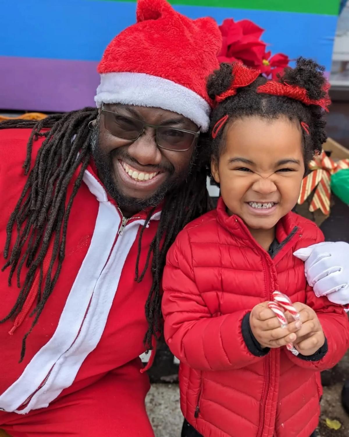 black santa smiles at the viewer while sitting next to a black toddler both are dressed in red