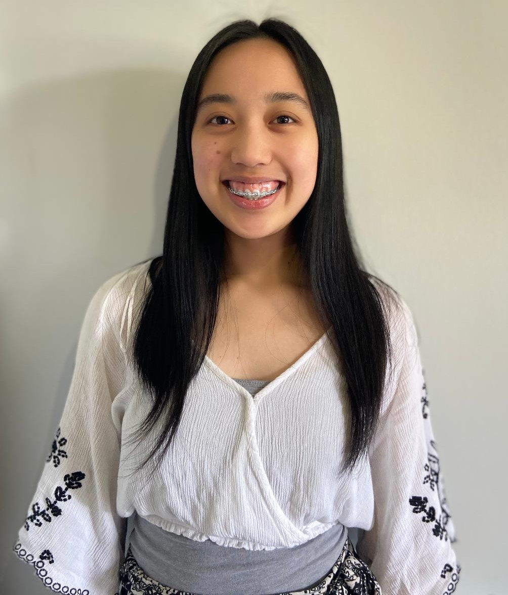 Light skinned female presenting person with long dark hair smiling brightly and wearing a white shirt with loose fitting sleeves that have dark embroidery on them
