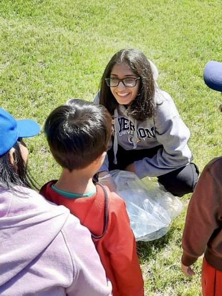 Female presenting person with dark shoulder length hair wearing glasses smiles at a child while squatting in an outdoor environment