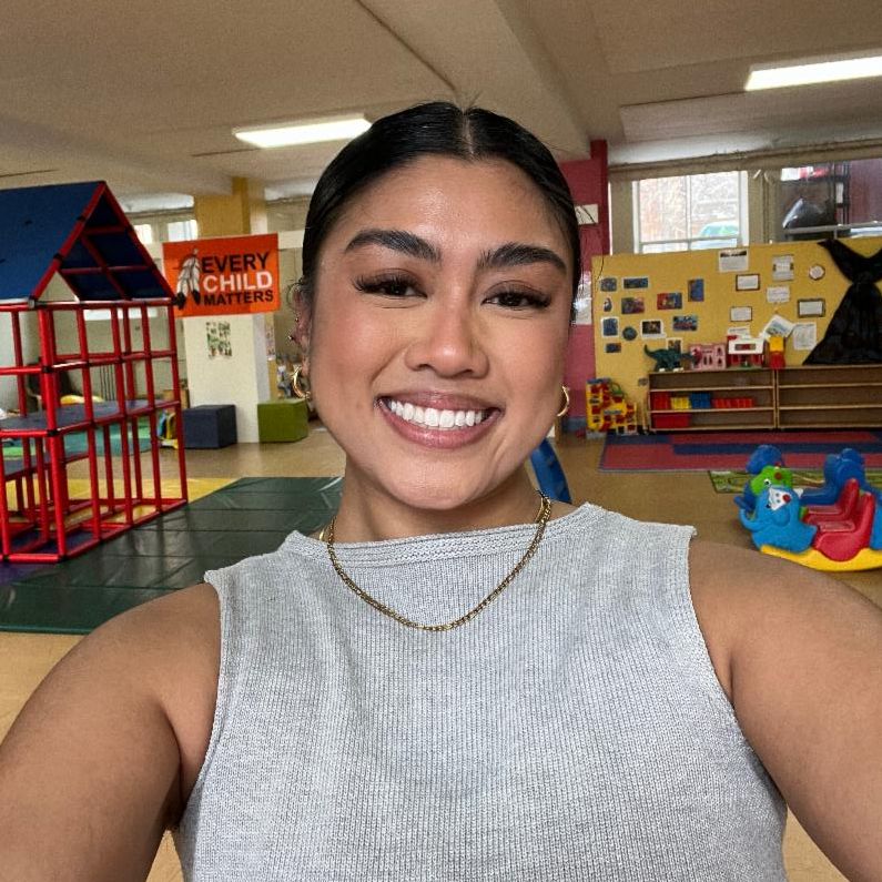 Female presenting person with light brown complexion and dark hair pulled back smiles brightly at the viewer in an indoor children's play setting