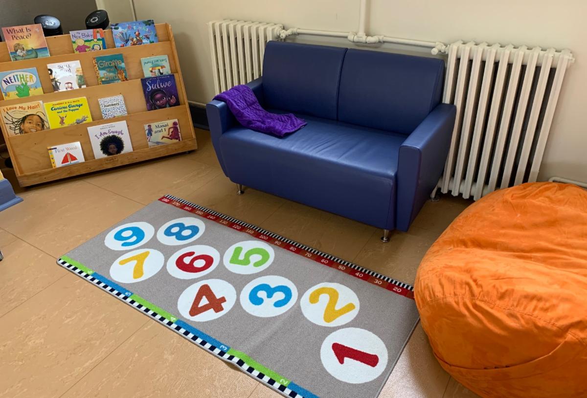 a children's reading area with a book display and a royal blue sofa and a very large orange bean bag seat with a mat on the floor with circles numbered 1 through 9