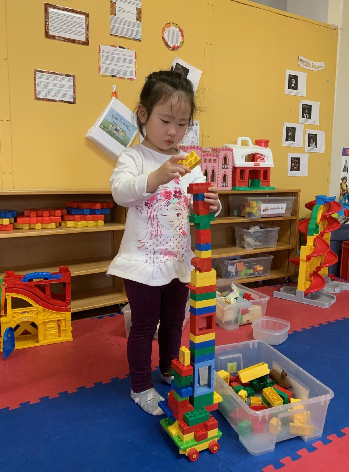 asian toddler stands while building a tower using coloured blocks while looking intently at the structure