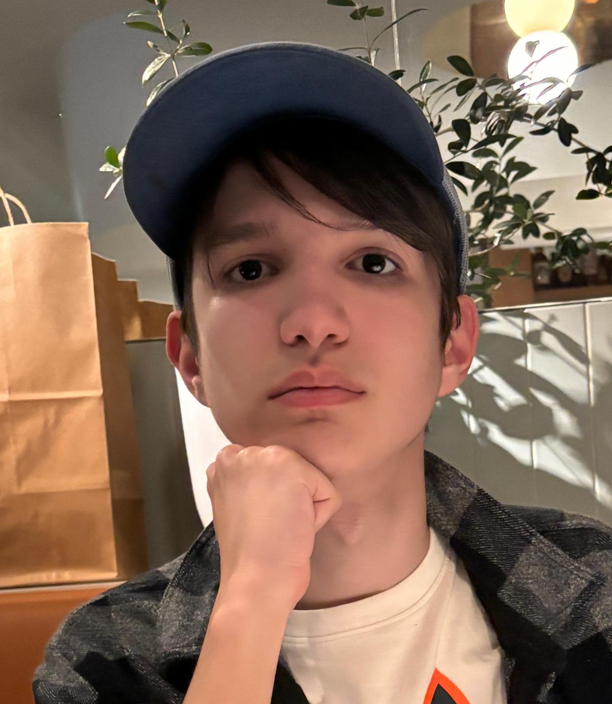 Headshot of  a male presenting youth wearing a ball cap with short dark hair and dark eyes gazes calmly at the viewer with a closed fist supporting his chin.