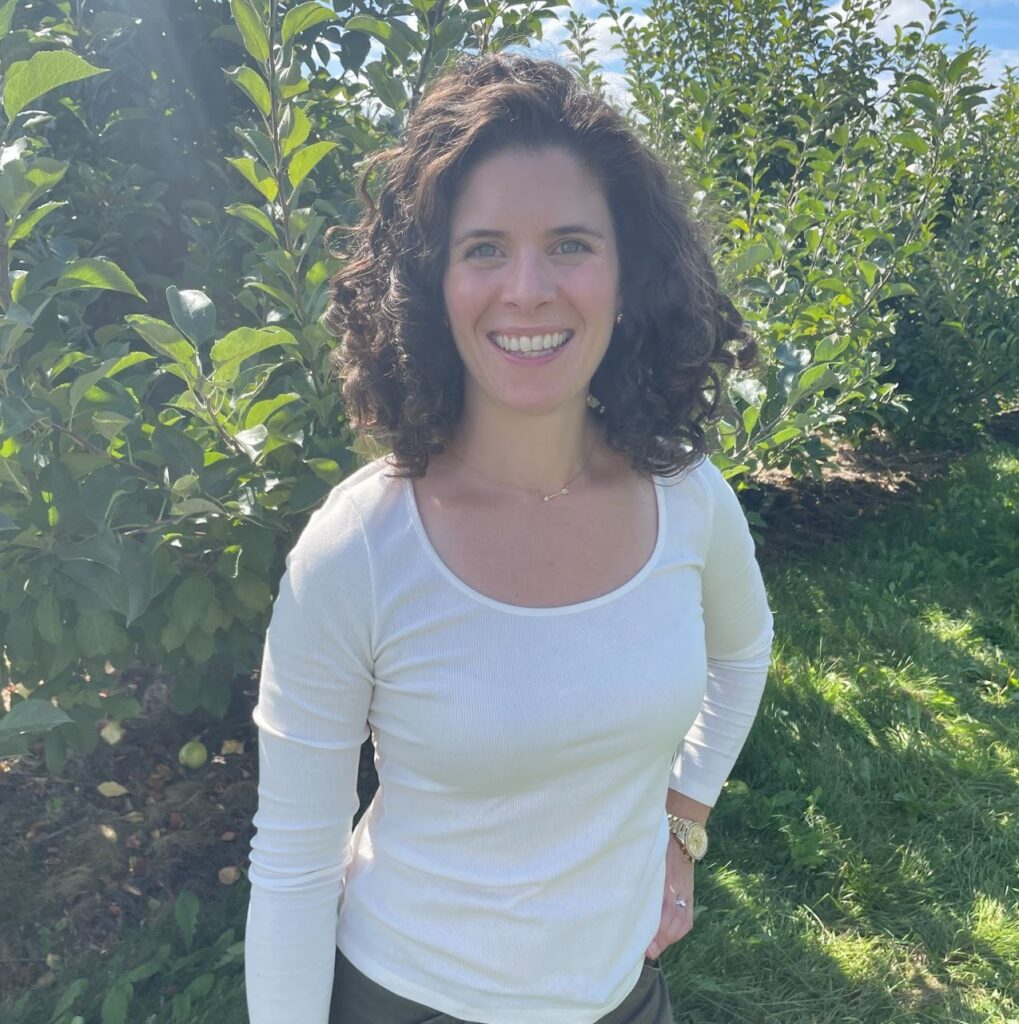 dark haired woman wearing a while long sleeved shirt smiles while standing in a leafy outdoor setting