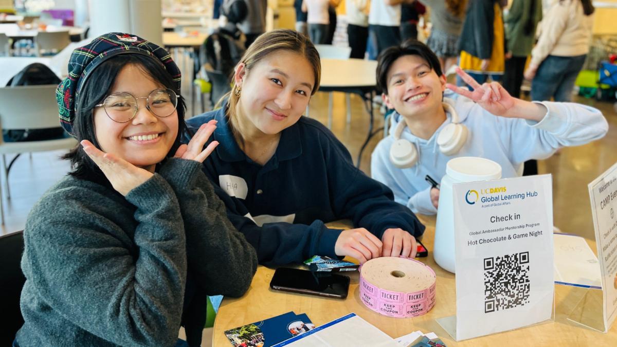 Three smiling students sit at a table during a UC Davis Global Ambassador Mentorship event with a check-in sign visible.