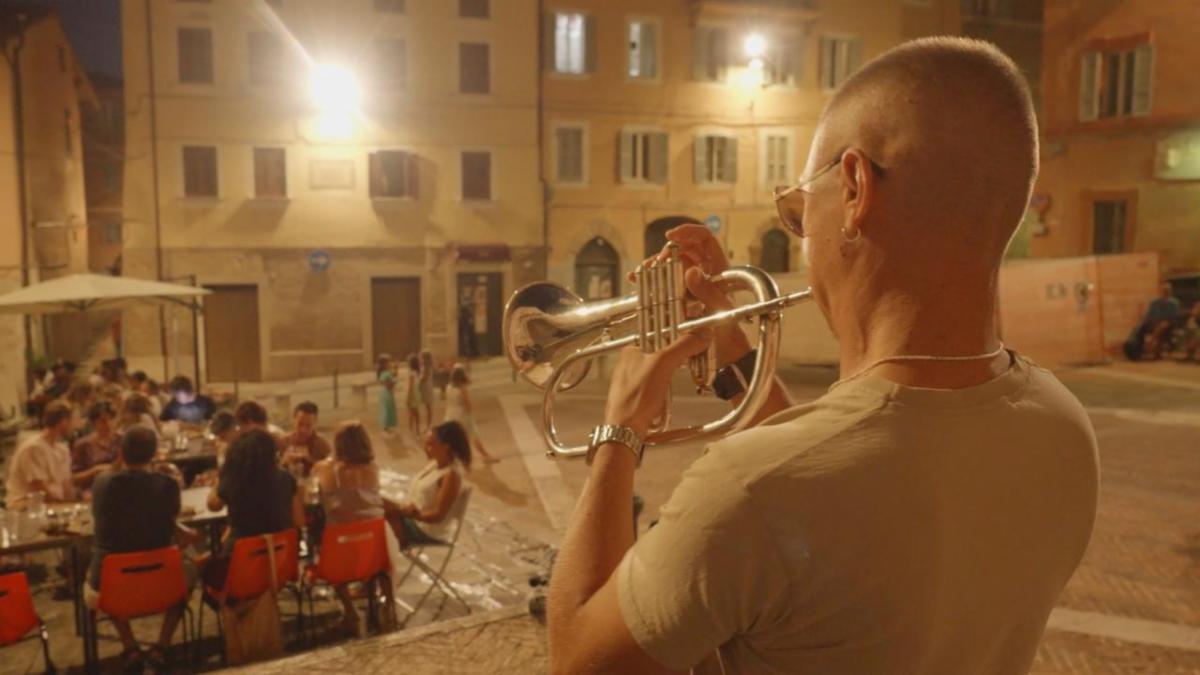 Giulio Angrisani plays the flugelhorn late at night in a square in Perugia outside the restaurant Il Garibaldino. Seen from behind, he performs beneath streetlights while diners sit at outdoor tables and historic buildings surround the square.  