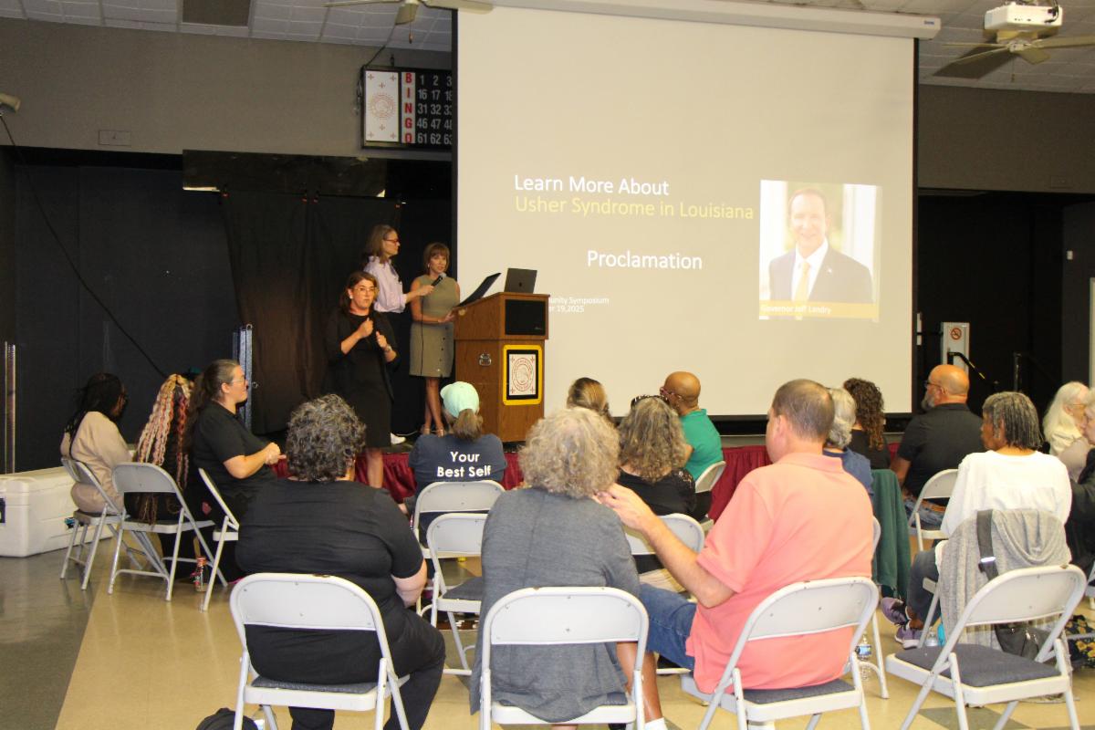 Picture is of Tillie Carrier GODA Senior Coordinator presenting the Governors proclamation at the Usher symposium on a stage to Dr. Lentz from LSU Nola health services a picture of Governor Landry is on a screen in the background and people are in chairs in the audience