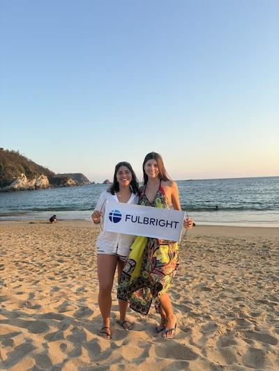 Two Fulbright grantees, including Daniella Passariello, stand on beach sand holding a sign that says, Fulbright