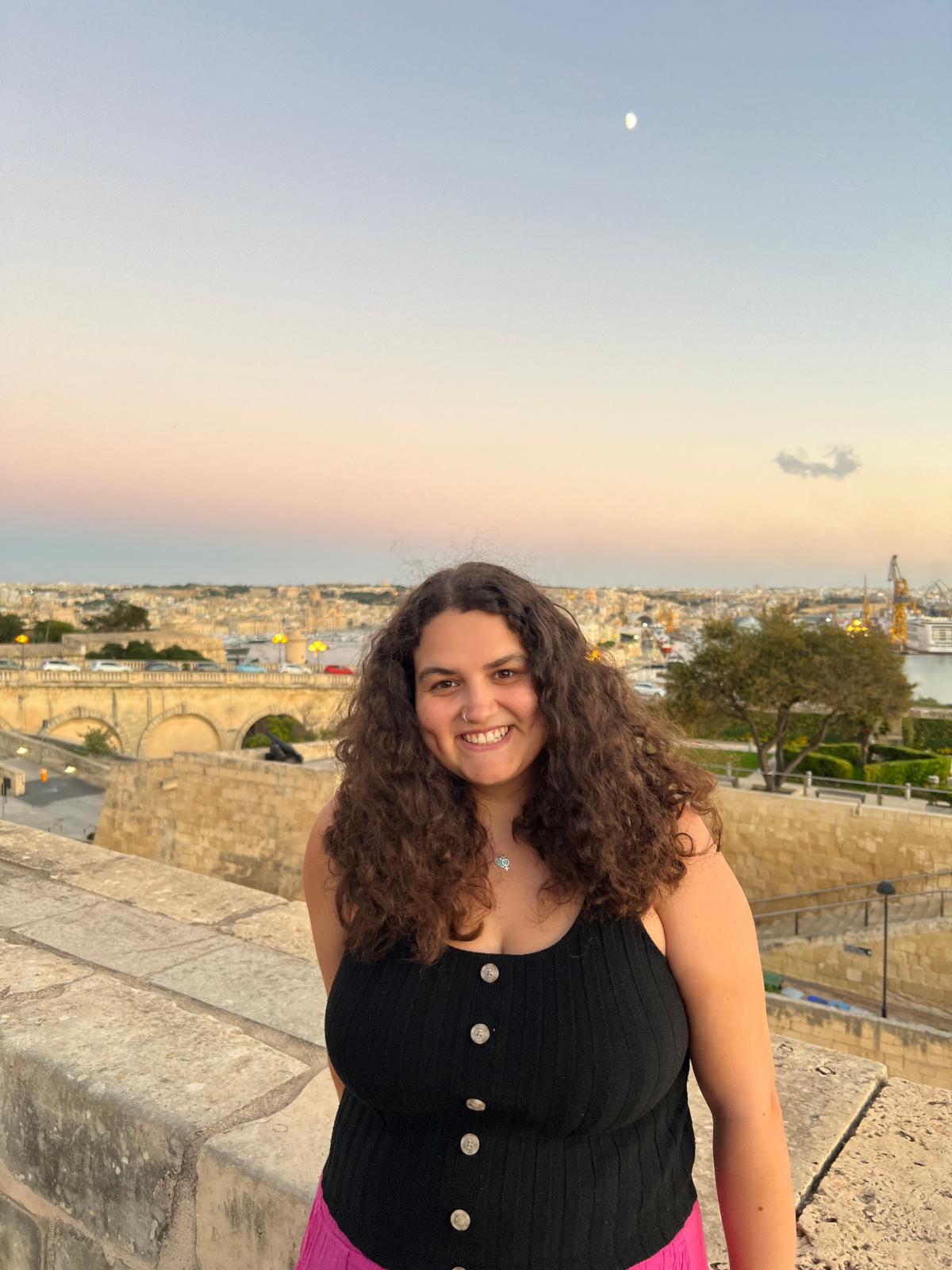 Kat Bouker poses in an outdoor setting with buildings and a pink and blue sky behind her