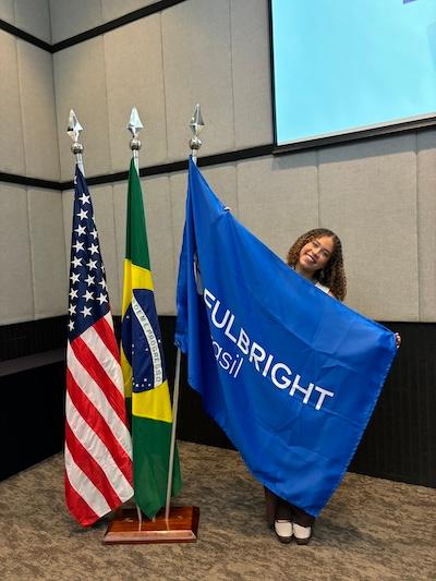 In an indoor setting, Maria Corvacho Marrugo holds a Fulbright Brasil flag next to the flags of Brazil and the United States