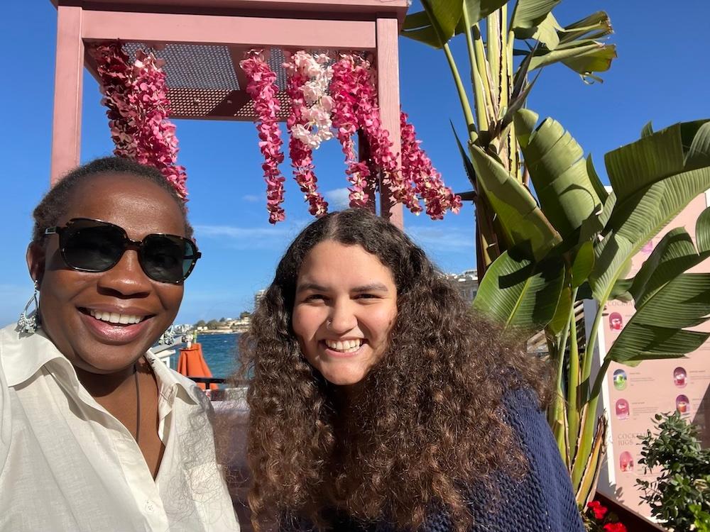 Dr. Charlene Brown-McKenzie and Kat Bouker pose together for a photo in an outdoor setting in front a foliage and a blue sky