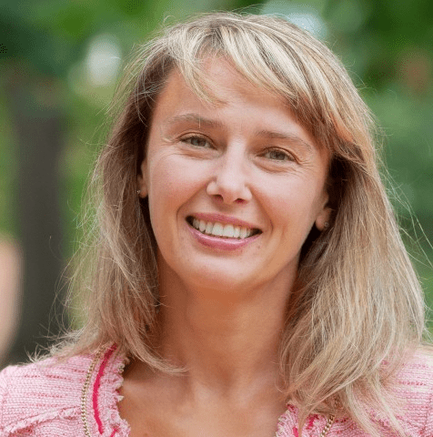 Headshot of a smiling woman with blonde hair, wearing a pink textured jacket, set against a soft-focus green outdoor background."