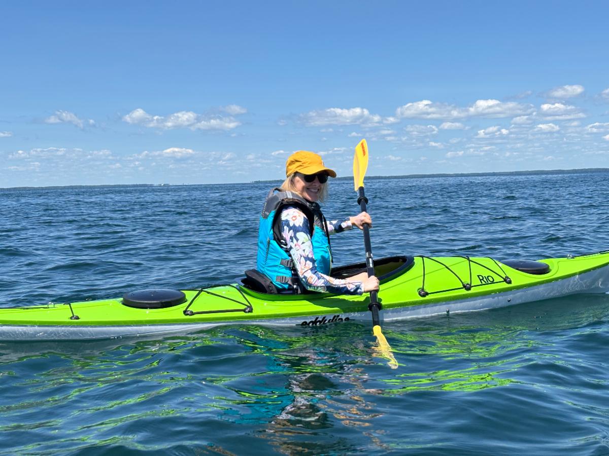 A person smiling while paddling a bright lime green kayak on calm, open blue water under a clear sunny sky.