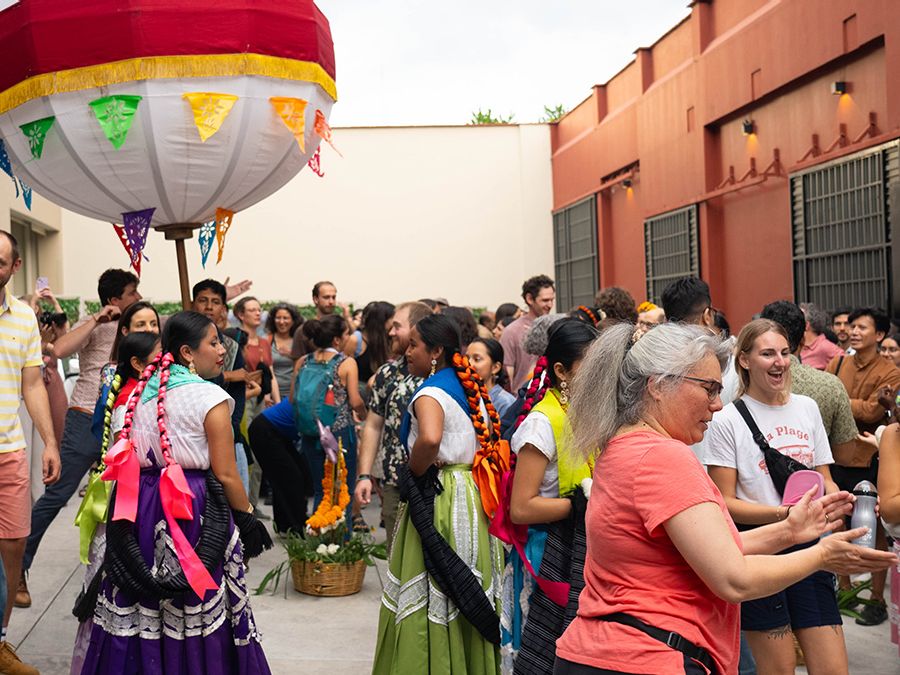A large, diverse group of people gathers in an outdoor courtyard in Oaxaca, Mexico, for a lively social event. In the foreground, two women in traditional Oaxacan dress—vibrant purple and green skirts with intricate white lace and ribbons braided into their hair—stand near a large, white and red decorative ceremonial balloon (marmota) adorned with colorful papel picado flags. Around them, many other attendees in casual attire are smiling, talking, and dancing. The background features warm, terracotta-colore