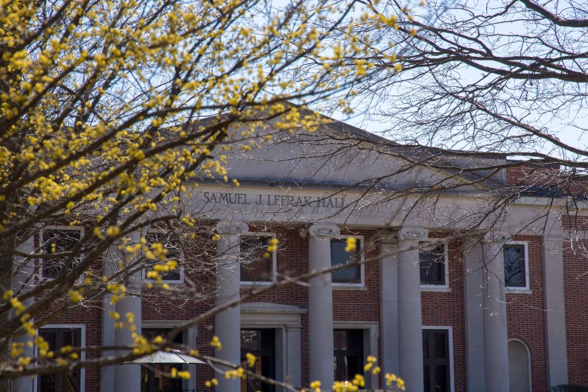 This image features a scenic view of LeFrak Hall at the University of Maryland, College Park, during a bright spring day. The red-brick building is showcased in its traditional Neoclassical style, with prominent white columns and a grand pediment partially framed by the vibrant yellow blossoms of a tree in the foreground. The sunlight highlights the building's historic facade, which houses various academic departments like Geography and Criminology, capturing a peaceful and scholarly atmosphere on the campus.
