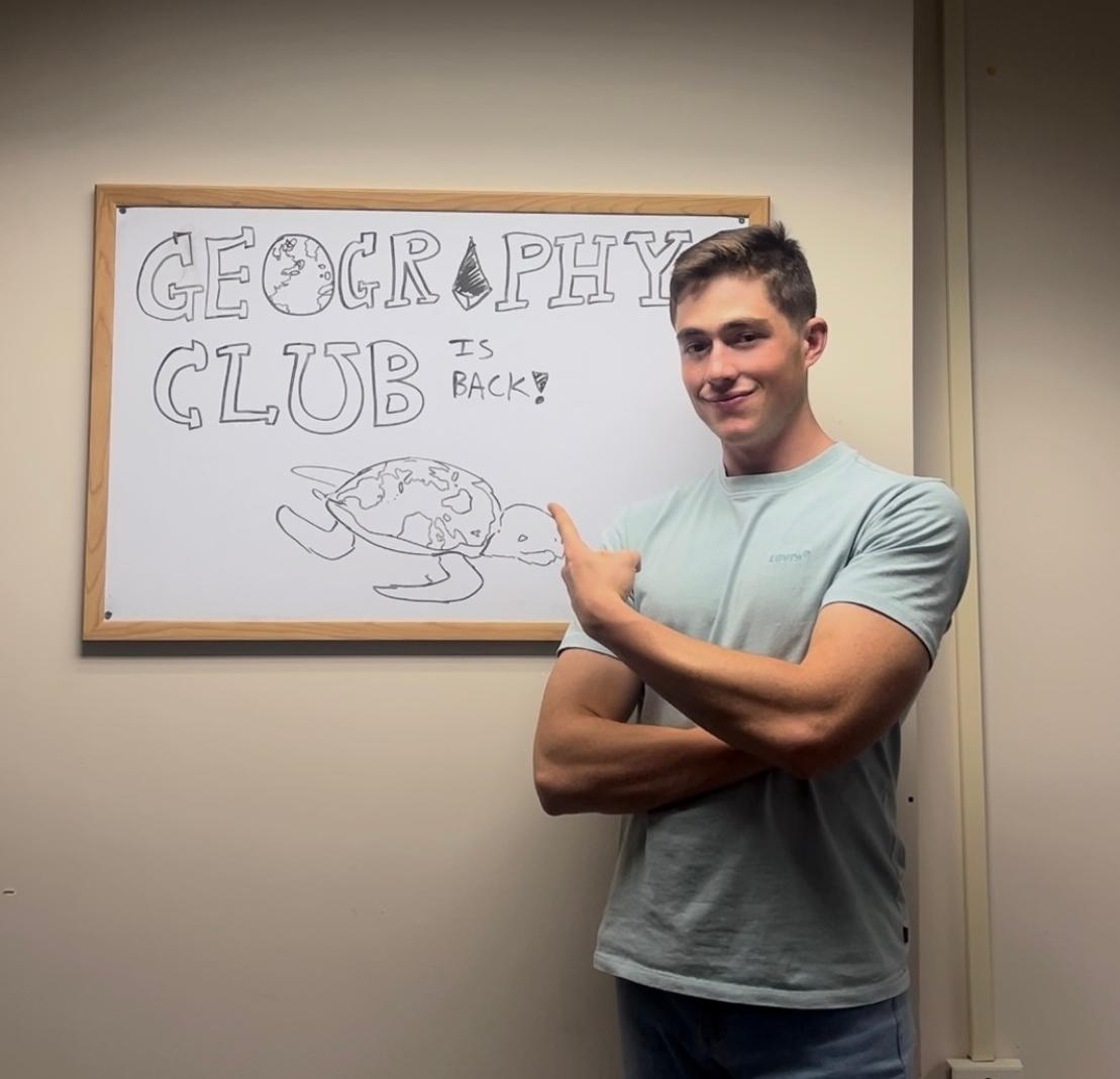 A young man in a light blue t-shirt stands next to a whiteboard framed on a plain wall. He has short brown hair, crossed arms, and points back over his shoulder toward the board with a slight smile. On the whiteboard, drawn in black marker, block letters spell out "GEOGRAPHY CLUB IS BACK!" with a globe integrated into the letter 'O' and an illustration of a sea turtle below the text whose shell resembles a world map.