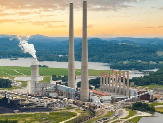 Aerial wide shot of a large coal-fired power plant nestled in a lush, mountainous valley. Two massive, slender concrete chimneys dominate the center, flanked by a shorter white cooling tower emitting a plume of steam. To the left, a large pile of coal sits near conveyor belts and rail lines, while a river curves around the facility in the foreground, all set against a backdrop of rolling green hills and a hazy blue mountain range under a warm, sunset-lit sky.