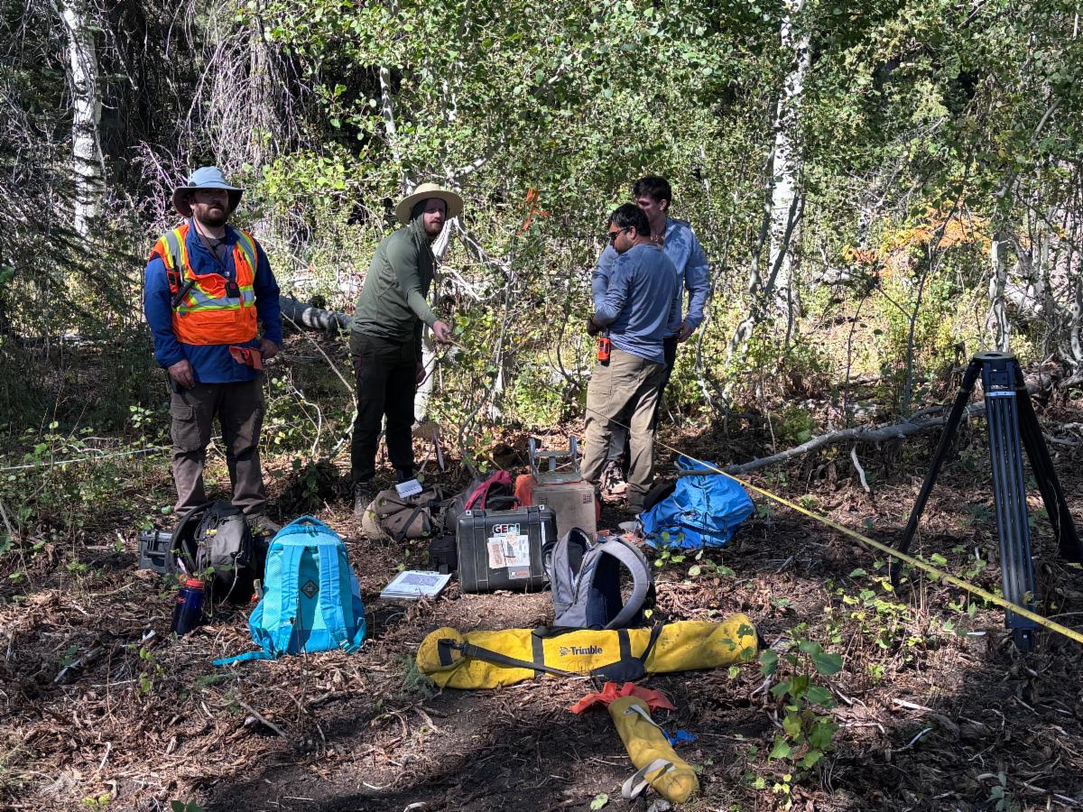 A field research team of four people gathered in a sunny, wooded clearing surrounded by aspen trees. On the left, a man wearing a high-visibility orange safety vest, a sun hat, and a backpack stands looking toward the camera. Next to him, another person in a wide-brimmed hat points toward something in the brush. To the right, two individuals stand near field equipment, including a tripod, measuring tape stretching across the ground, and various gear bags, backpacks, and hard cases labeled with Trimble and GEO logos on the forest floor.