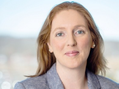 A professional headshot of a woman with strawberry-blonde hair and blue eyes, smiling warmly while wearing a blue textured blazer and pearl earrings against a soft, blurred outdoor background.