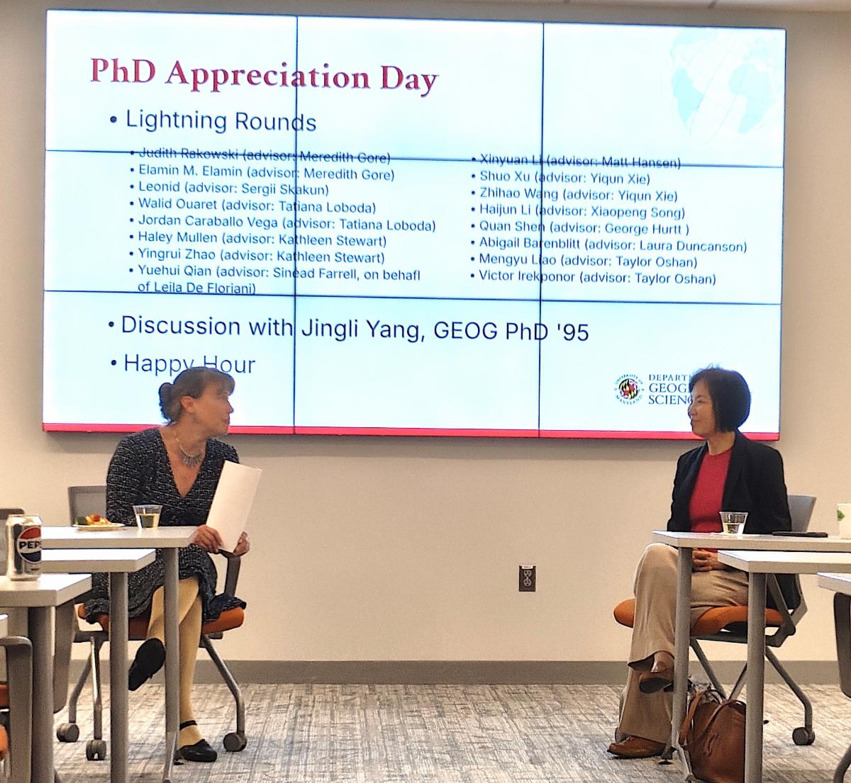 A professor in a dark patterned dress and a woman in a black blazer and pink top sit in chairs facing each other, engaged in a discussion. Behind them, a large digital screen displays PhD Appreciation Day.