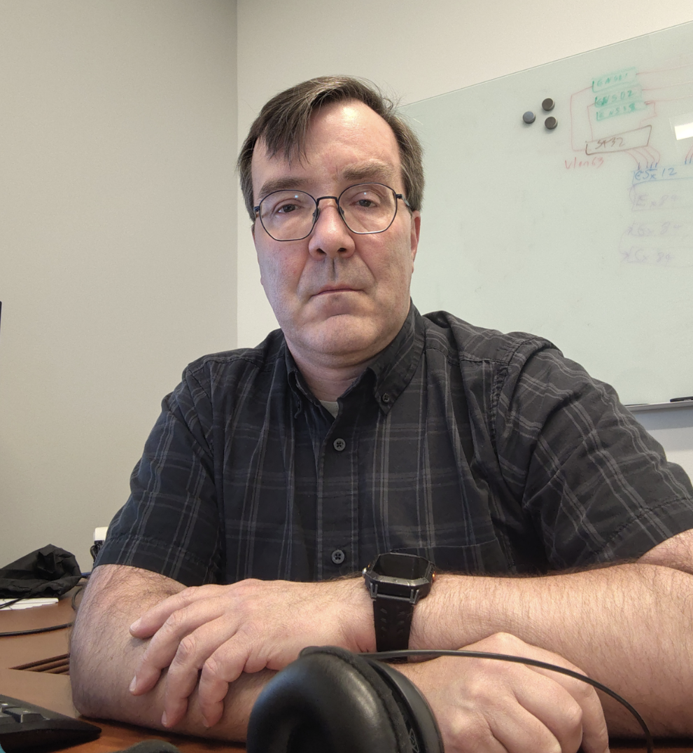 Close-up portrait of a man with short brown hair and round glasses, wearing a dark plaid button-up shirt and a black smartwatch. He is sitting at a wooden desk with his arms crossed, looking forward with a neutral expression.