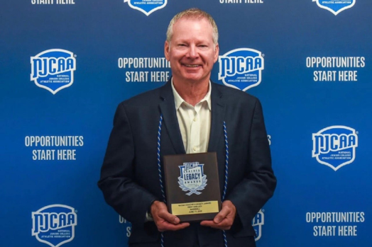 Kent Shelley smiles in front of an NJCAA banner with an award plaque in his hands.