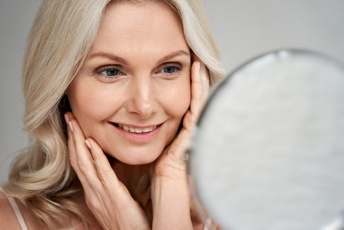 Happy 50s middle aged woman model touching face skin looking in mirror. Smiling mature older lady pampering_ enjoying healthy skin care_ aging beauty_ skincare treatment cosmetic products concept.