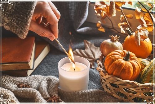 Hand with burning match lighting a candle on the windowsill with cozy autumn still life with pumpkins_ knitted woolen sweater and books. Autumn home decor. Cozy fall mood. Thanksgiving. Halloween.