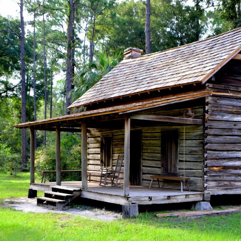 Weathered log cabin with a shingled roof, covered porch, rocking chair, and surrounding trees.