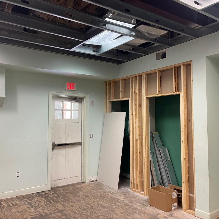 Renovation area with an illuminated EXIT sign, exposed wall framing, dismantled ceiling, and construction materials. Transcribed Text:  EXIT