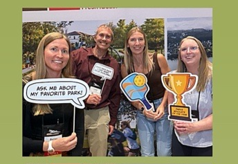 Four adults pose at a park-themed photobooth, holding speech-bubble, sign, illustrated, and trophy props.
