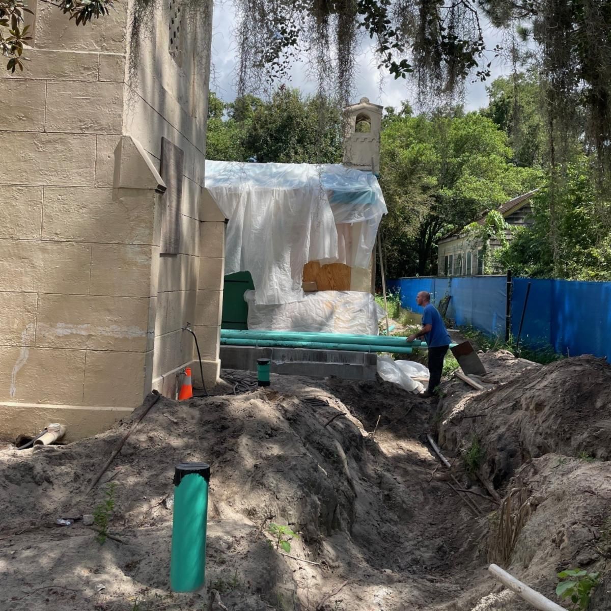 Construction work beside a building, with trenches, green pipes, plastic sheeting, and a worker beneath trees draped with Spanish moss.