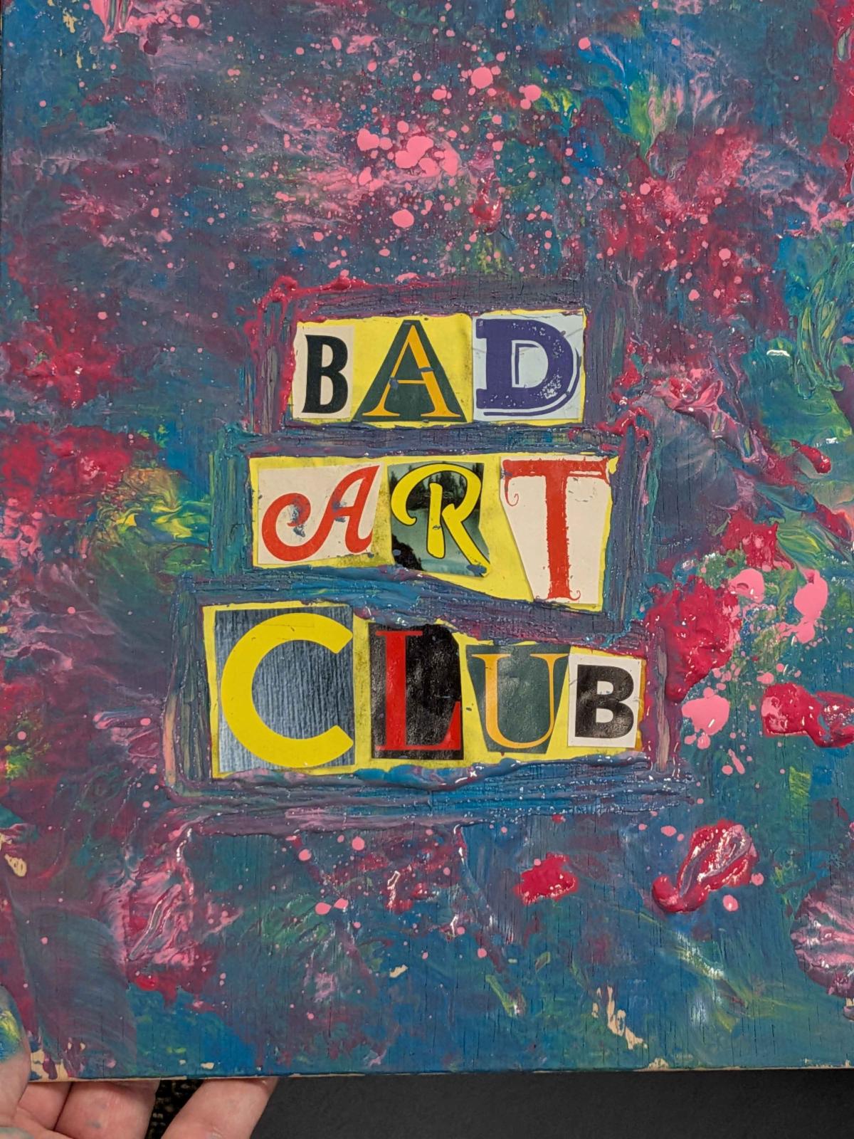 A hand holding a paint-splattered canvas. "Bad art club" is spelled on it with letters from different magazines.