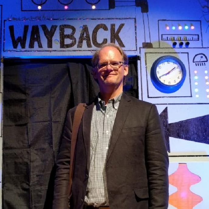 A man with glasses, wearing khaki pants and blazer, stands in front of a cardboard machine labeled "Wayback"