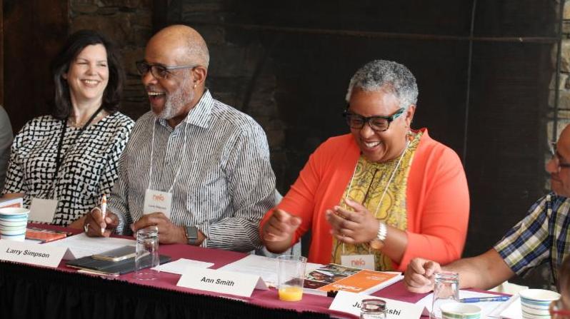 A black man and a black woman sitting at a conference table share a laugh.