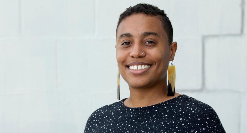 A queer woman of color in long earrings and a starry printed shirt smiles at the camera