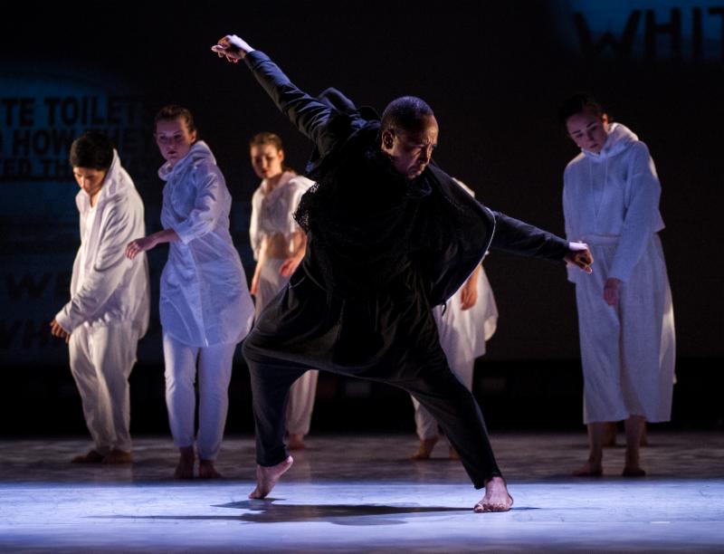 A man in black dances in front of a line of dancers in all white