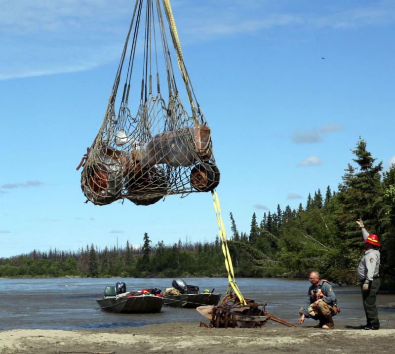 Image of the Last Homesteader's tractor being airlifted out by helicopter. Trees and a river are visible in the background as a ranger points upward.