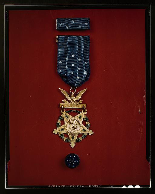 Image of a Medal of Honor, the highest military decoration the U.S. government awards. Golden colored medal with a blue ribbon on a red background.