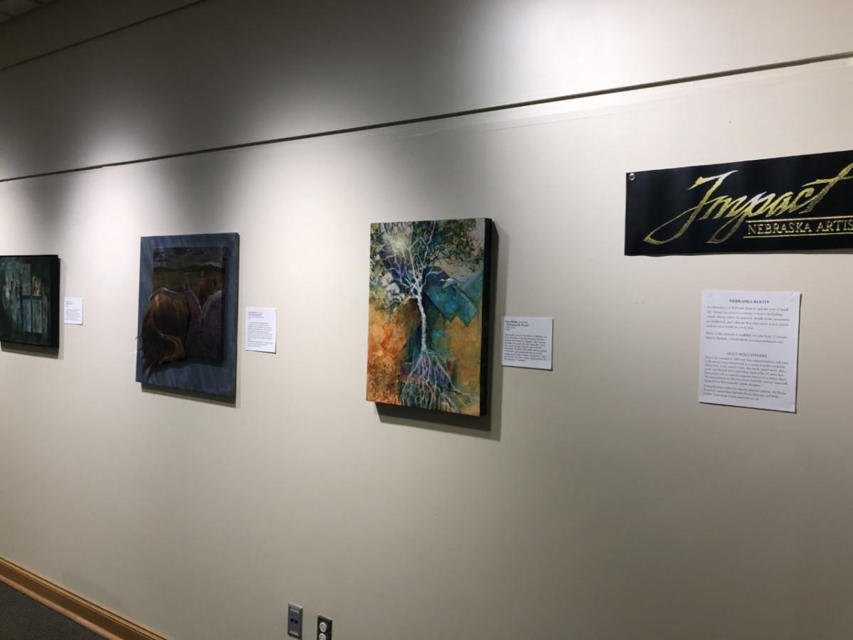 Impact: Nebraska Artists, Nebraska Roots exhibit at the Homestead Heritage Center. Several pieces of art hanging on the wall.