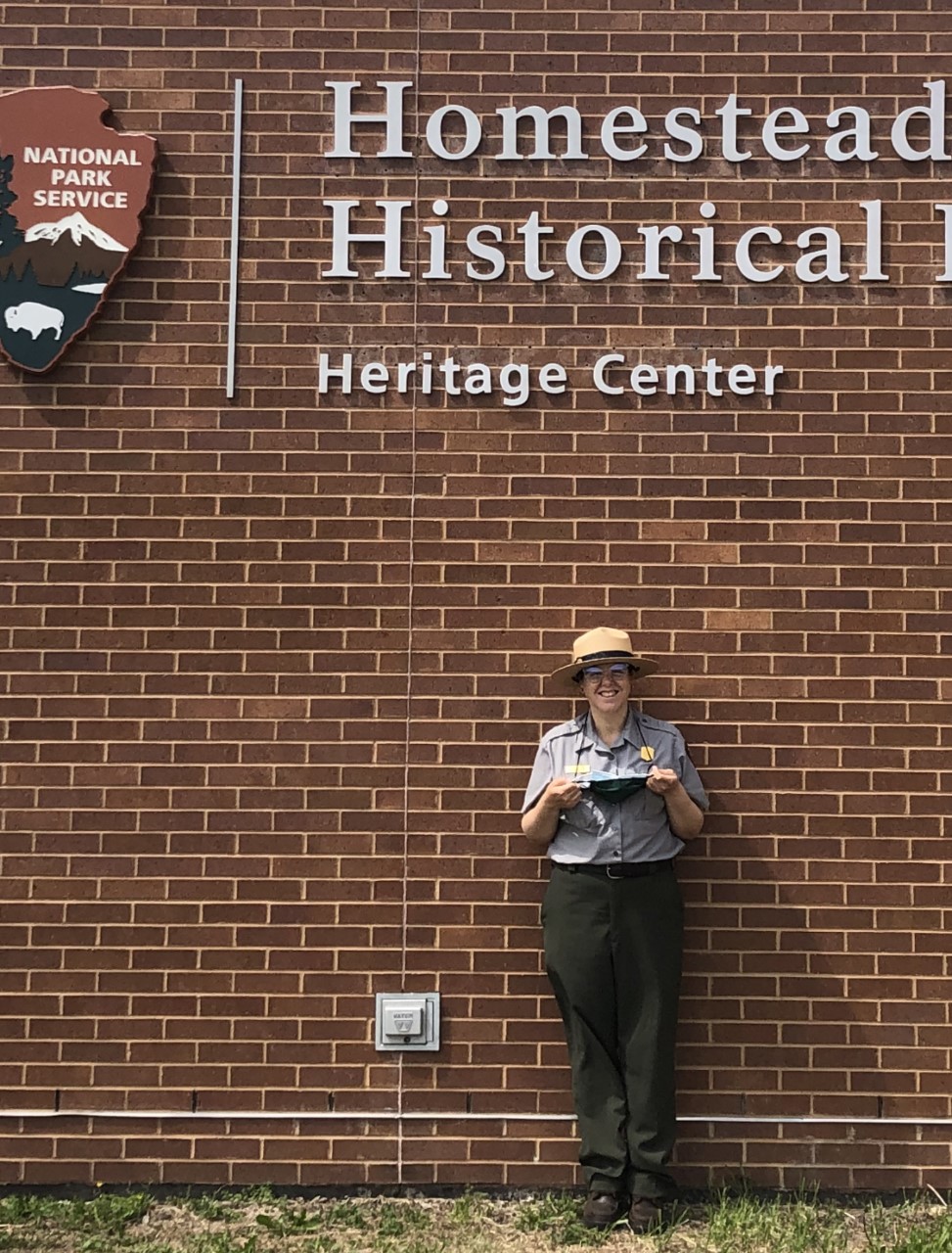 Homestead Education Tech Liliana Valderrama standing in front of the Homestead Heritage Center. A brick wall and sign are visible in the background.