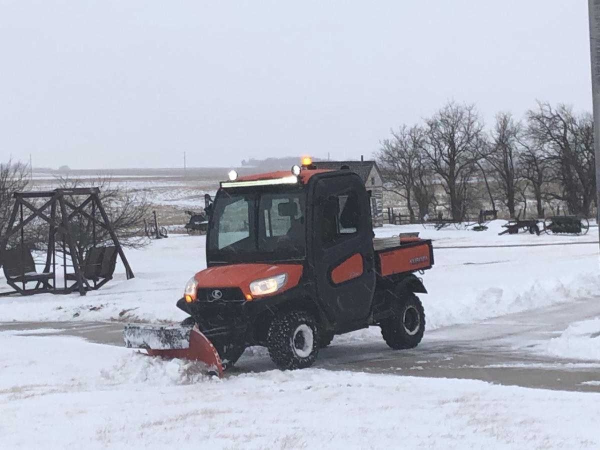 An orange Utility Task Vehicle plows snow from the grounds of Homestead National Historical Park. Trees and a cabin are visible in the background.